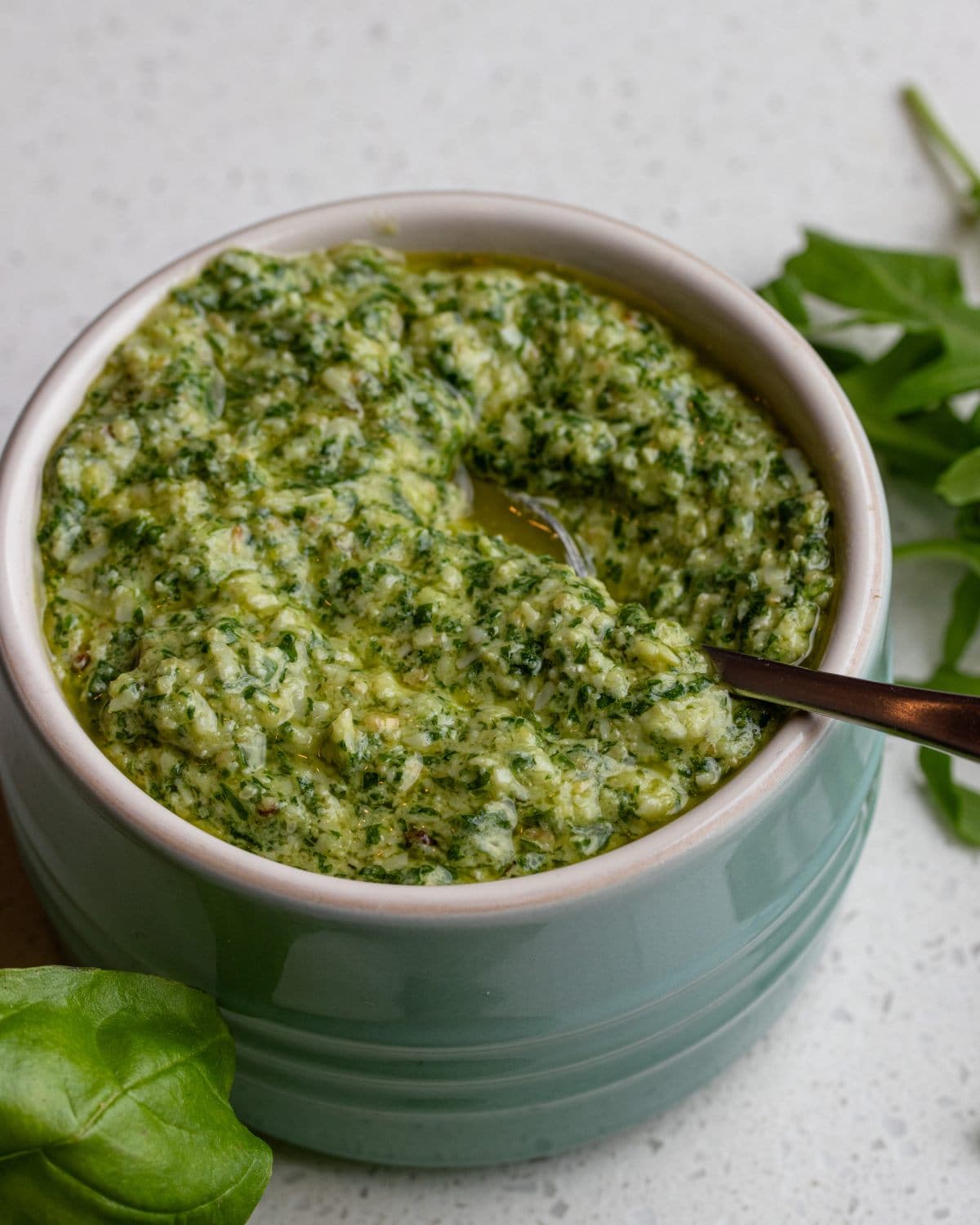 A ramekin of arugula pesto on a bench top. Teaspoon resting in with fresh basil and arugula leaves around it.