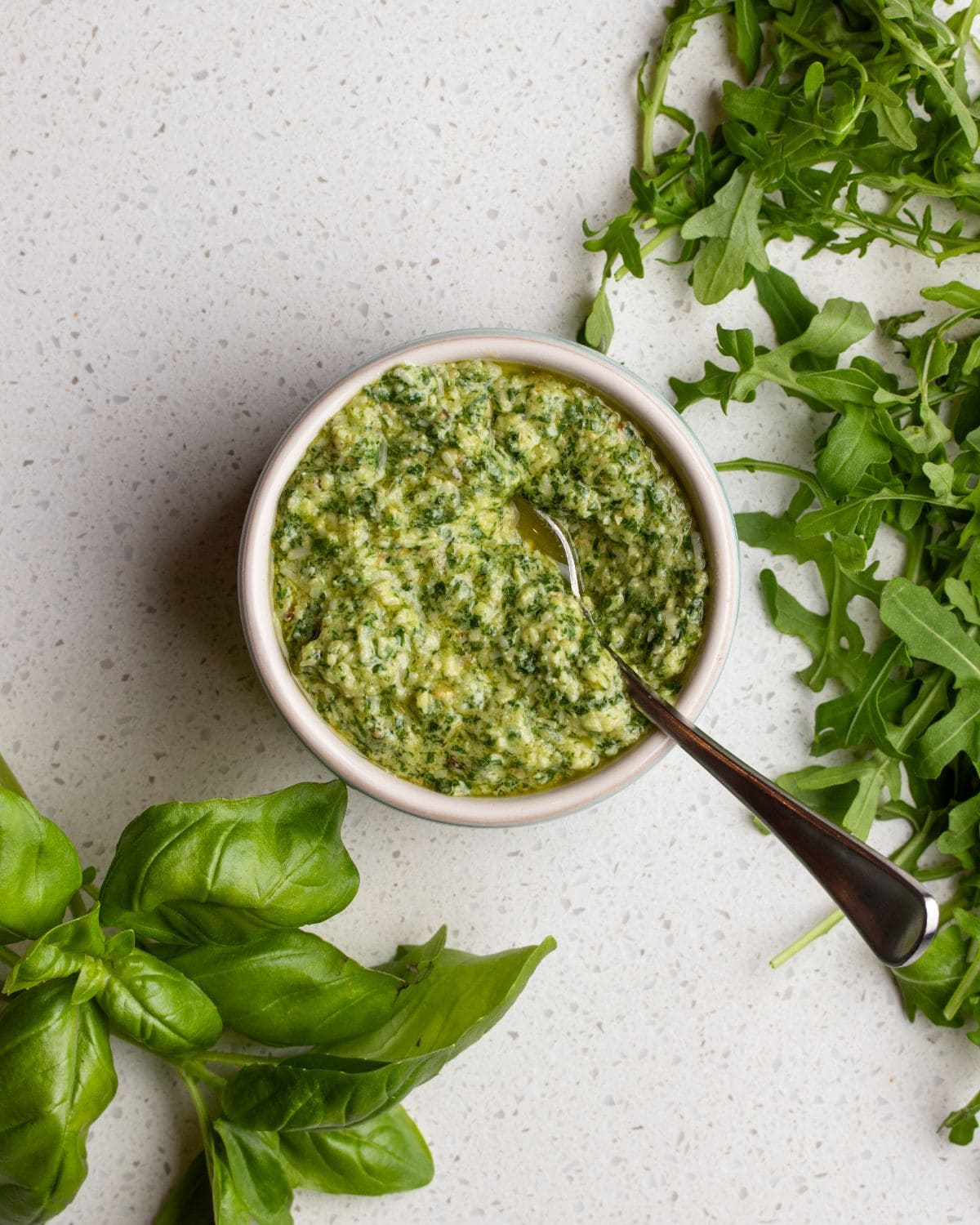 A ramekin of arugula pesto on a bench top. Teaspoon resting in with fresh basil and arugula leaves around it.