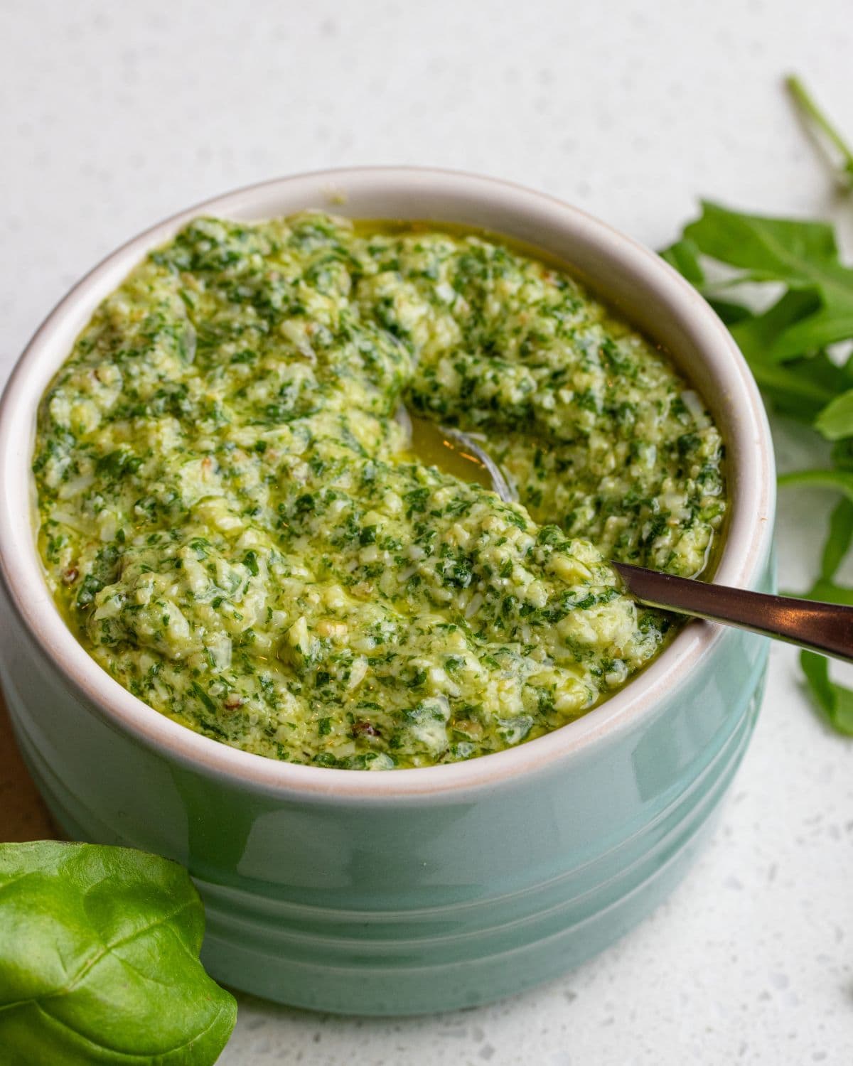 A ramekin of arugula pesto on a bench top. Teaspoon resting in with fresh basil and arugula leaves around it.