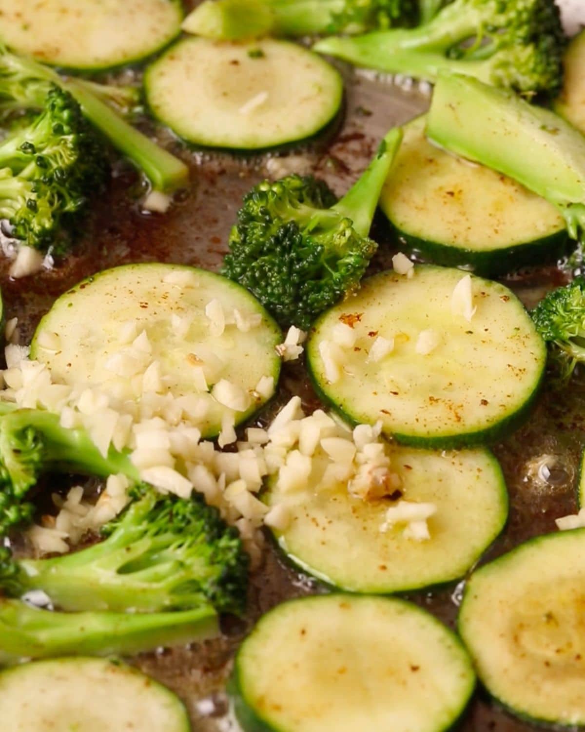Chopped zucchini and broccoli frying in a pan with chopped garlic on top.