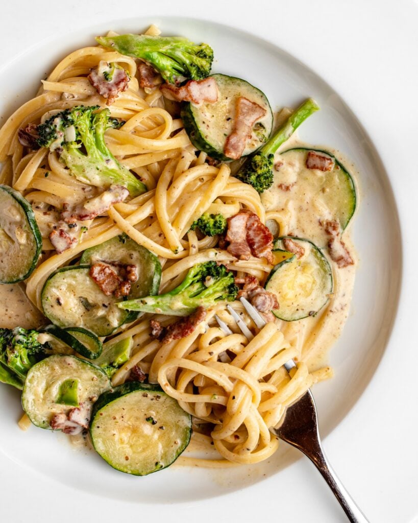 Cooked spaghetti in a creamy bacon pasta sauce on a white plate. Broccoli and courgette slices mived in with pasta twirled around the fork.