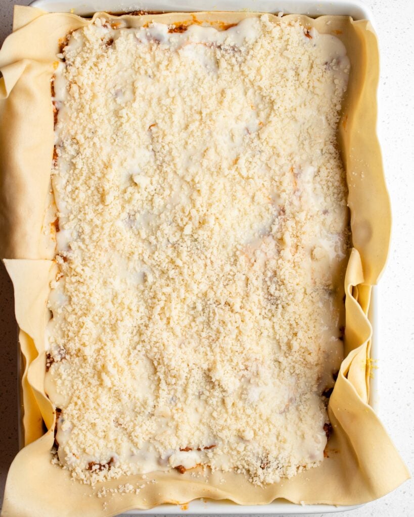 Final assembled lasagne ready to be baked. Top has layer of parmesan cheese and the pasta sheets are overhanging the sides of the dish.