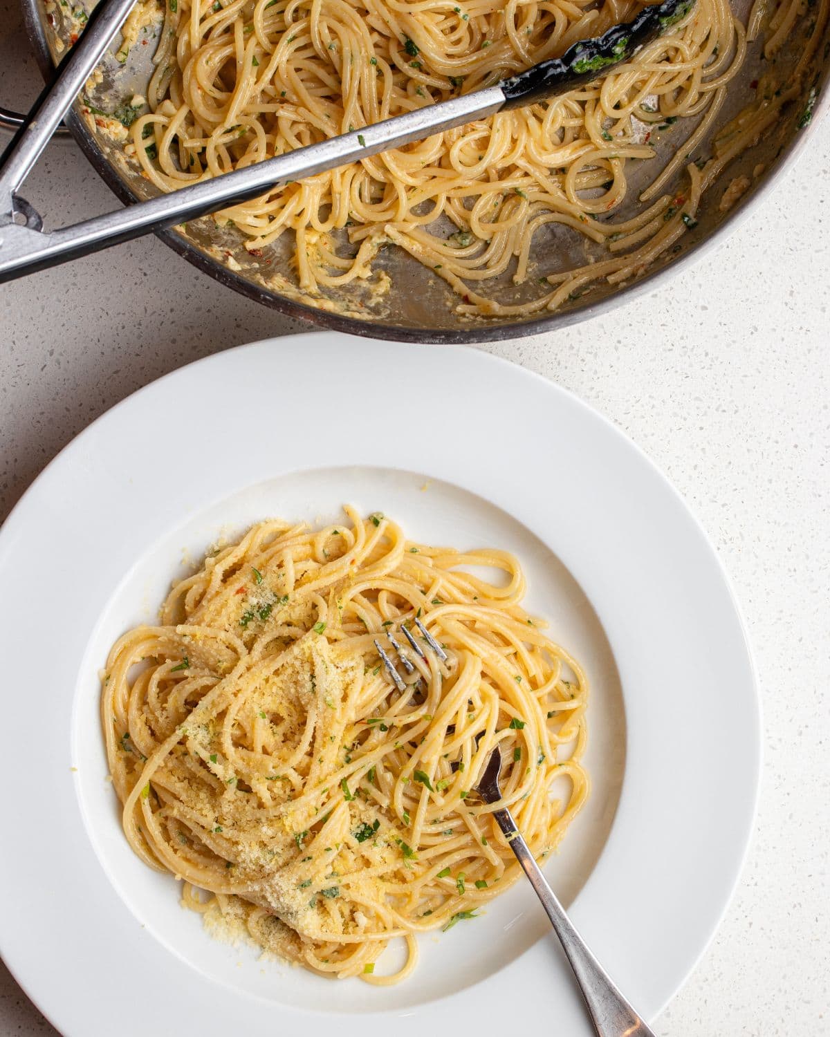 Bench top with white plate with the lemon butter spaghetti. Skillet above with remaining pasta.