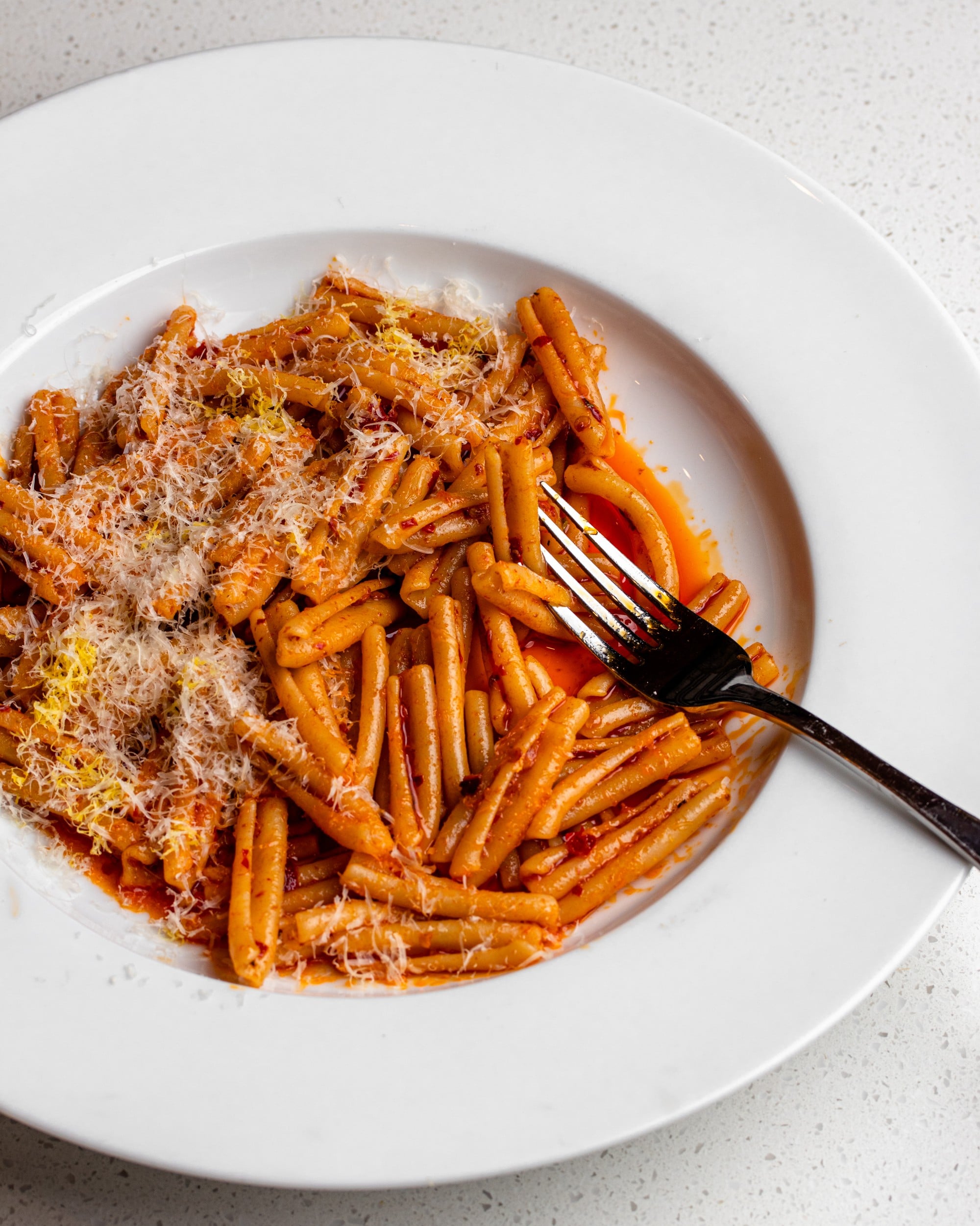 A white plate on a bench with cooked pasta in a red nduja butter sauce. Freshly grated parmesan and lemon zest on top and a fork resting into pasta on right.