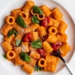 White plate with rigatoni in a roasted tomato sauce. Torn bail leaves on top and a fork resting.