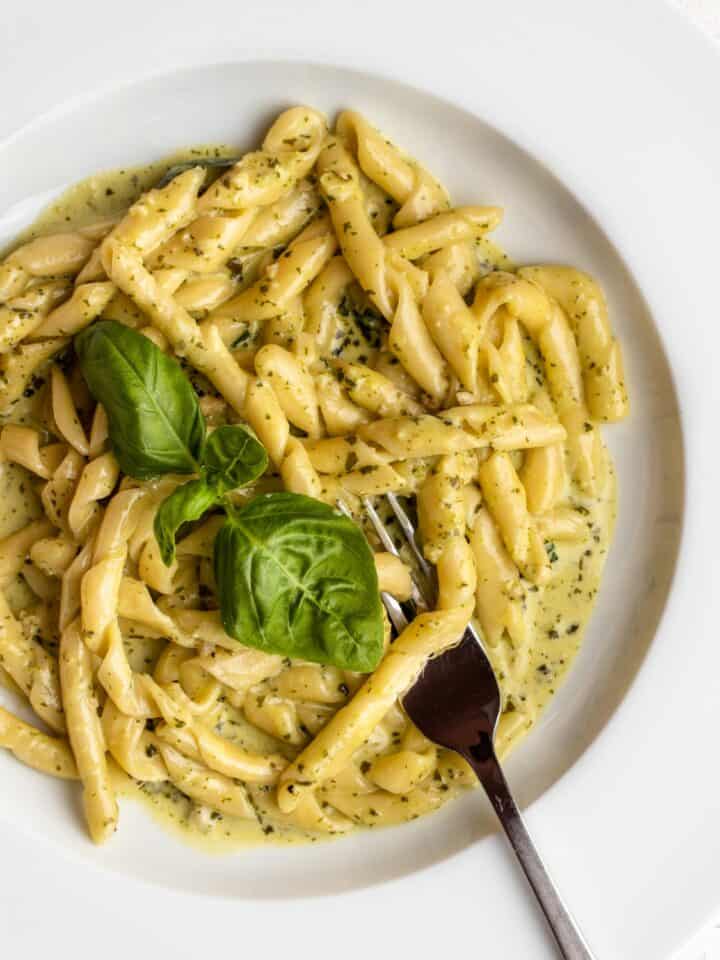 White plate with busiate pasta in a creamy pesto sauce. Fresh basil leaves on top and a fork resting in.