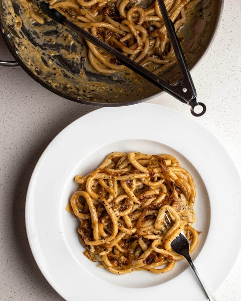 Pici pasta in a cacio e pepe sauce with golden caramelized onions mixed through. A fork testing on plate with noodles twirled around it. Fry pan seen above with the rest.