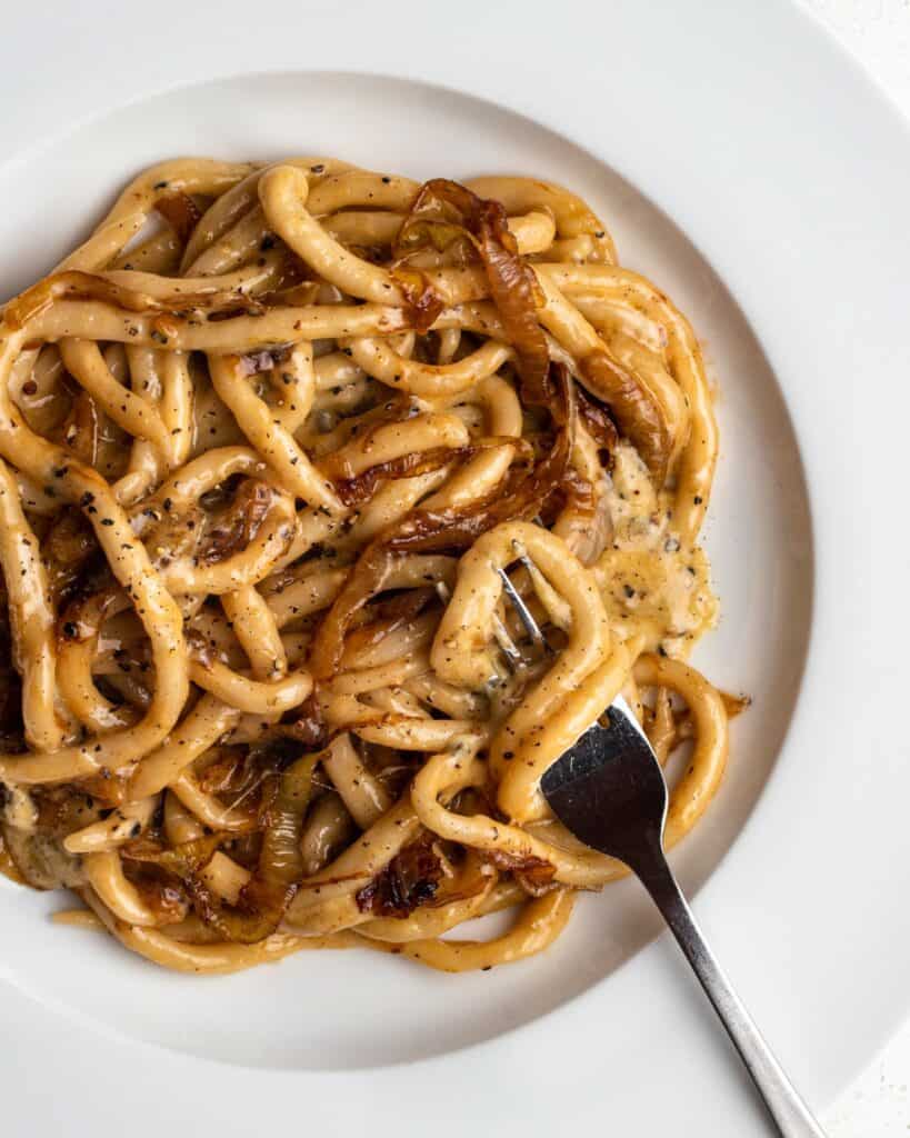 Pici pasta in a cacio e pepe sauce with golden caramelized onions mixed through. A fork testing on plate with noodles twirled around it.