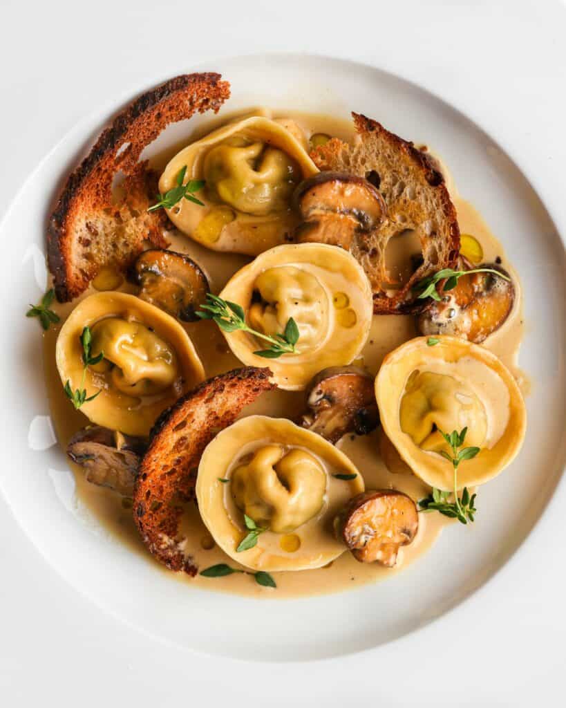 White plate with cappelletti in a cream sauce. Mushrooms, sourdough croutons and fresh thyme scattered.