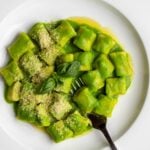 White plate with gnocchi in a vibrant green pea sauce. Fresh basil and parmesan on top. Fork resting into gnocchi.