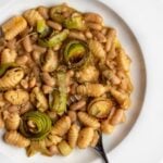 A white plate with gnocchi pasta in a miso sauce with cannellini beans, golden brown leek rounds and fresh rosemary on top. Fork resting into the pasta.