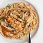 Fettuccine in a creamy sauce with golden brown sliced chicken on top. Fresh rosemary leaves and lemon zest sprinkled on top.