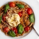 White plate with fettuccine in a buttery cherry tomato sauce. Fresh basil, grated parmesan cheese and a torn ball of burrata on top.