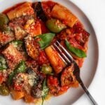 White plate with rigatoni pasta in a red tomato sauce with eggplant cubes, olives and fresh basil. A fork resting in.