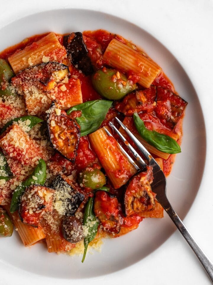 White plate with rigatoni pasta in a red tomato sauce with eggplant cubes, olives and fresh basil. A fork resting in.