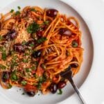 White plate with spaghetti in a puttanesca sauce. Fork with twirled noodles around it and fresh parsley and parmesan on top.