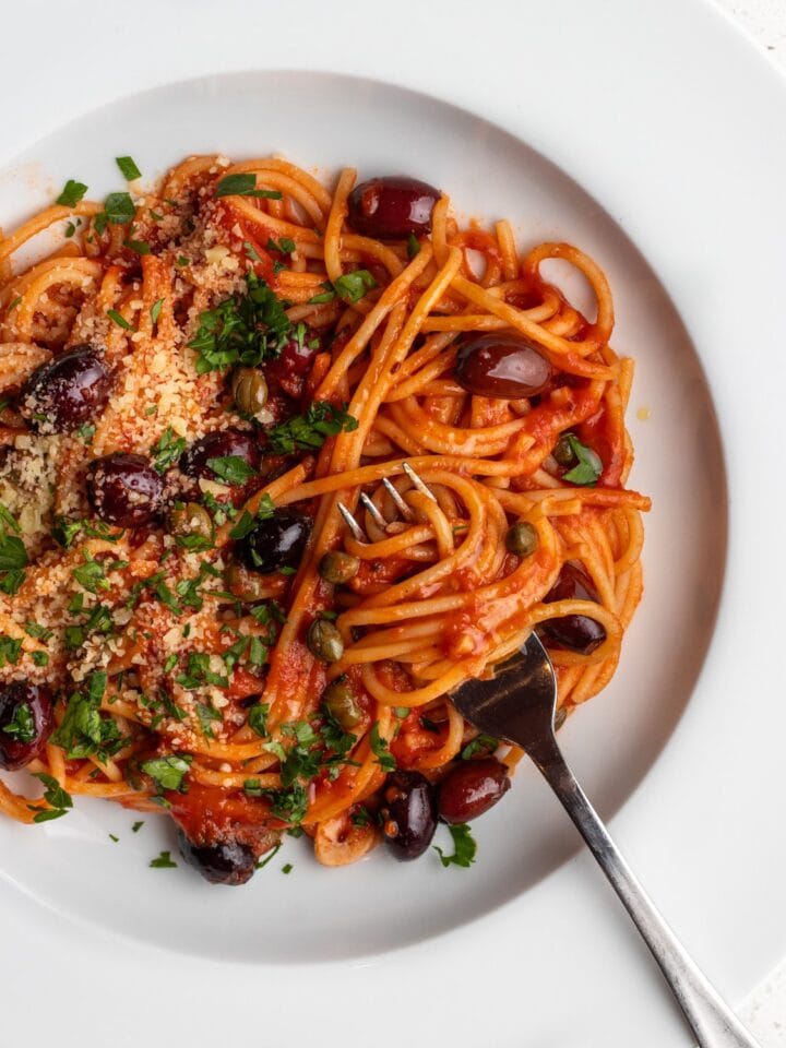 White plate with spaghetti in a puttanesca sauce. Fork with twirled noodles around it and fresh parsley and parmesan on top.