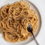 White plate with spaghetti in a cacio e pepe sauce. Fresh cracked black pepper on top and a fork resting into noodles.