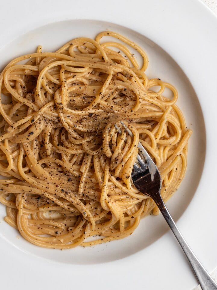 White plate with spaghetti in a cacio e pepe sauce. Fresh cracked black pepper on top and a fork resting into noodles.