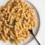 White plate with fusilli pasta in a creamy miso sauce. Fresh rosemary scattered on top and a fork resting in.