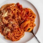 White plate with spaghetti in a red amatriciana sauce. Grated parmesan cheese and crispy gianciale pieces on top. Fork resting.