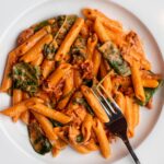 White plate with penne pasta in a creamy sun-dried tomato sauce. Fork resting in.