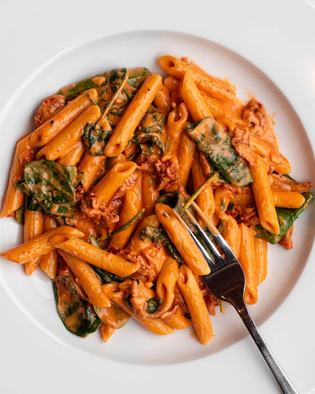 White plate with penne pasta in a creamy sun-dried tomato sauce. Fork resting in.