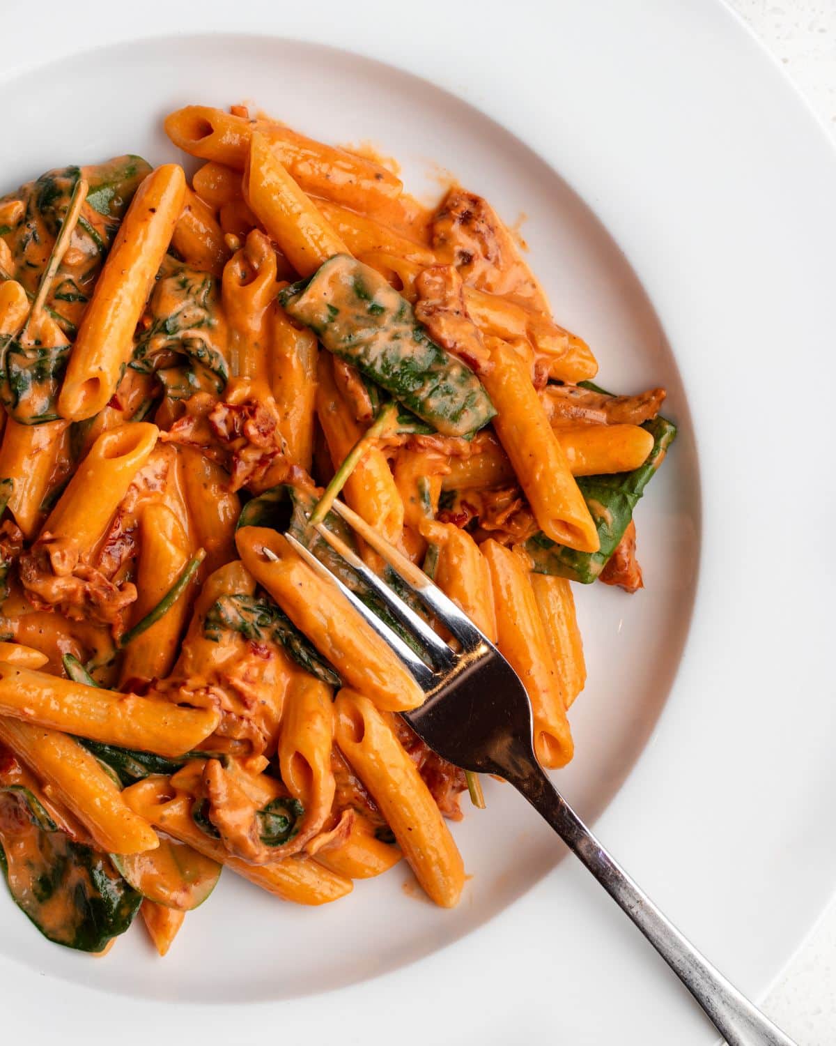 Penne pasta in a creamy sun-dried tomato sauce. Fork resting in and wilted baby spinach on top.