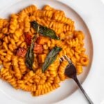 White plate with fusilli pasta in a creamy butternut sauce. Crispy sage leaves on top and a fork resting in.