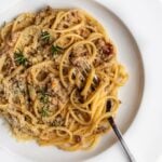 White plate with spaghetti in a white bolognese sauce. Grated parmesan and fresh rosemary leaves on top and a fork twirling spaghetti resting.