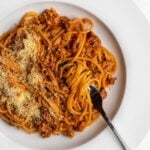 White plate with spaghetti bolognese. Grated parmesan on top and a fork resting.