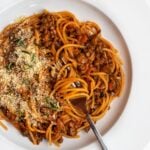 White plate with spaghetti in a vegetarian bolognese sauce. Freshly grated parmesan and fresh rosemary on top.