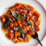 Crispy sweet potato gnocchi on a white plate with a brown butter sauce and crispy sage leaves. Fork resting in.