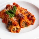 Three stuffed cannelloni on a white plate with grated parmesan cheese and fresh basil on top.
