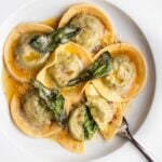 White plate with round ravioli, a foamy butter sauce and crisp sage. A fork digging in to a ravioli to show green spinach filling.