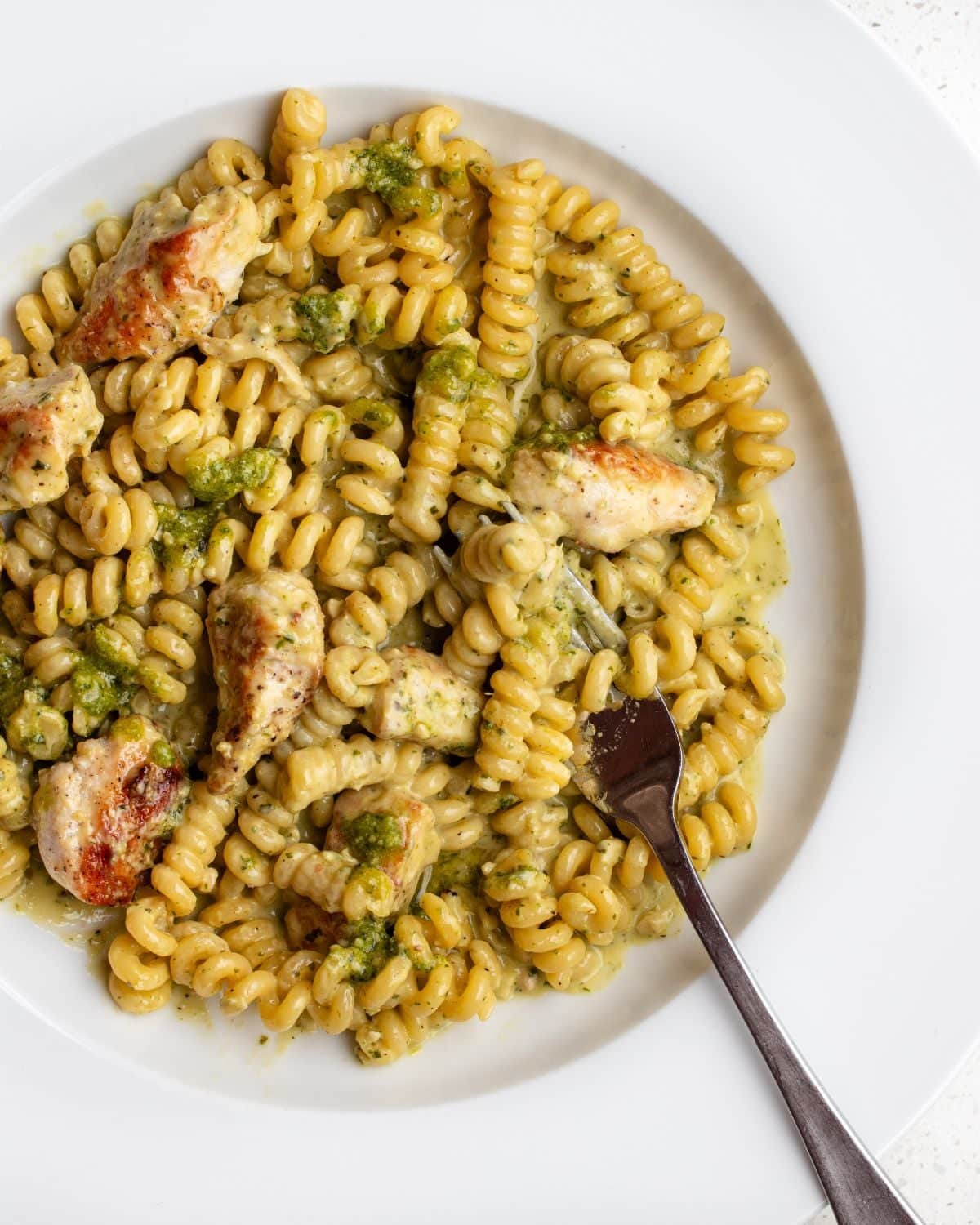 White plate with fusilli pasta in a creamy pesto sauce with chicken. Fork resting in.