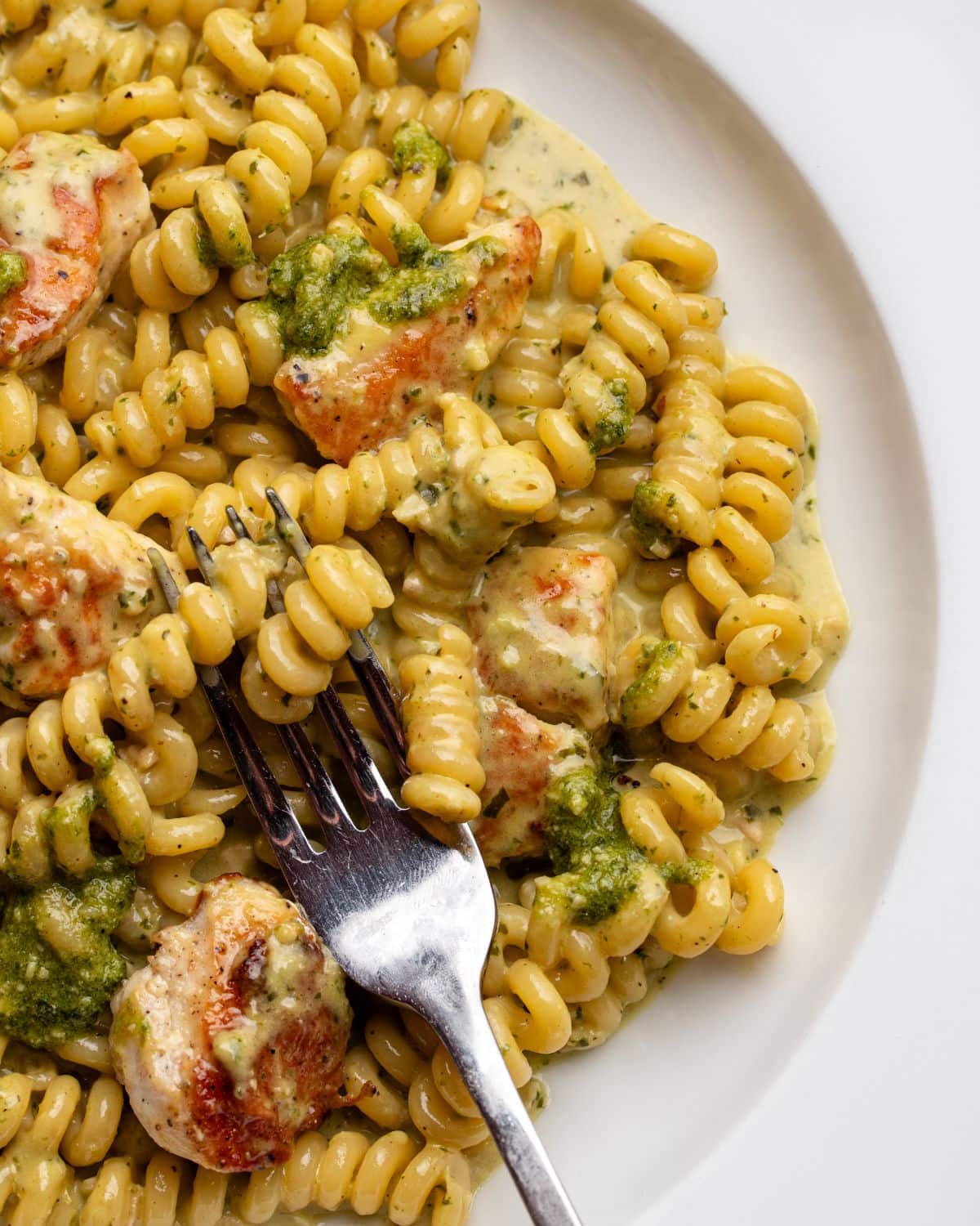 White plate with fusilli pasta in a creamy pesto sauce with chicken. Fork resting in.