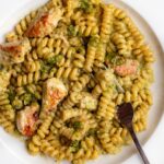 White plate with fusilli pasta in a creamy pesto sauce with chicken. Fork resting in.