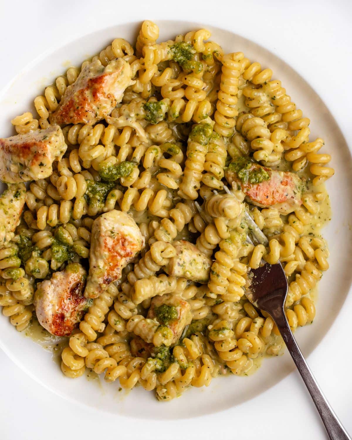 White plate with fusilli pasta in a creamy pesto sauce with chicken. Fork resting in.
