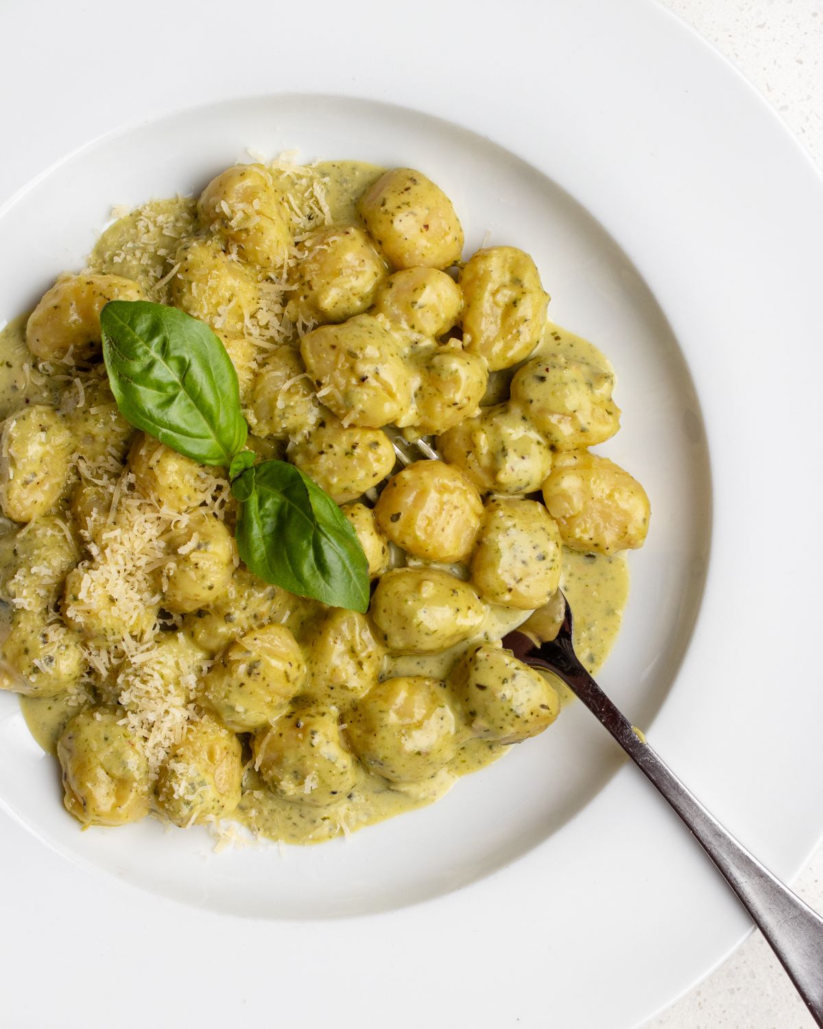 White plate with gnocchi in a creamy pesto sauce. Basil leaves on top and a fork resting in.