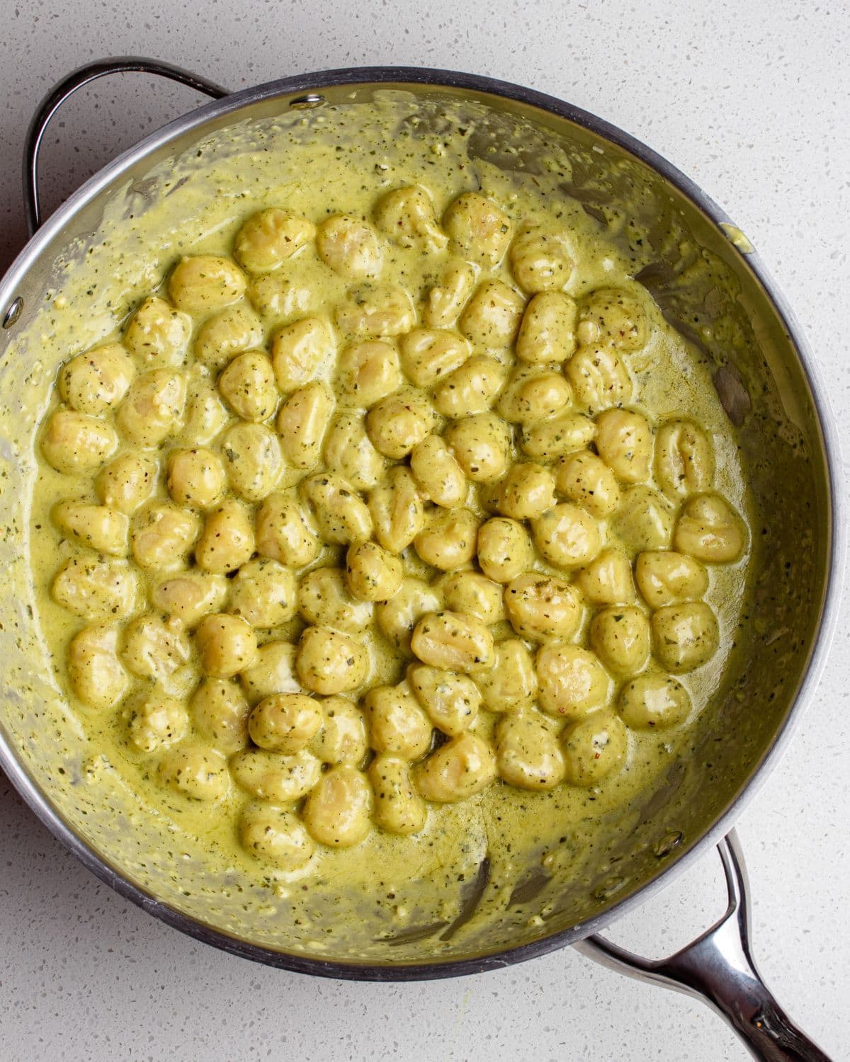 Gnocchi in a pesto sauce in a stainless steel fry pan on a white bench top.