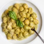 White plate with gnocchi in a creamy pesto sauce. Basil leaves on top and a fork resting in.