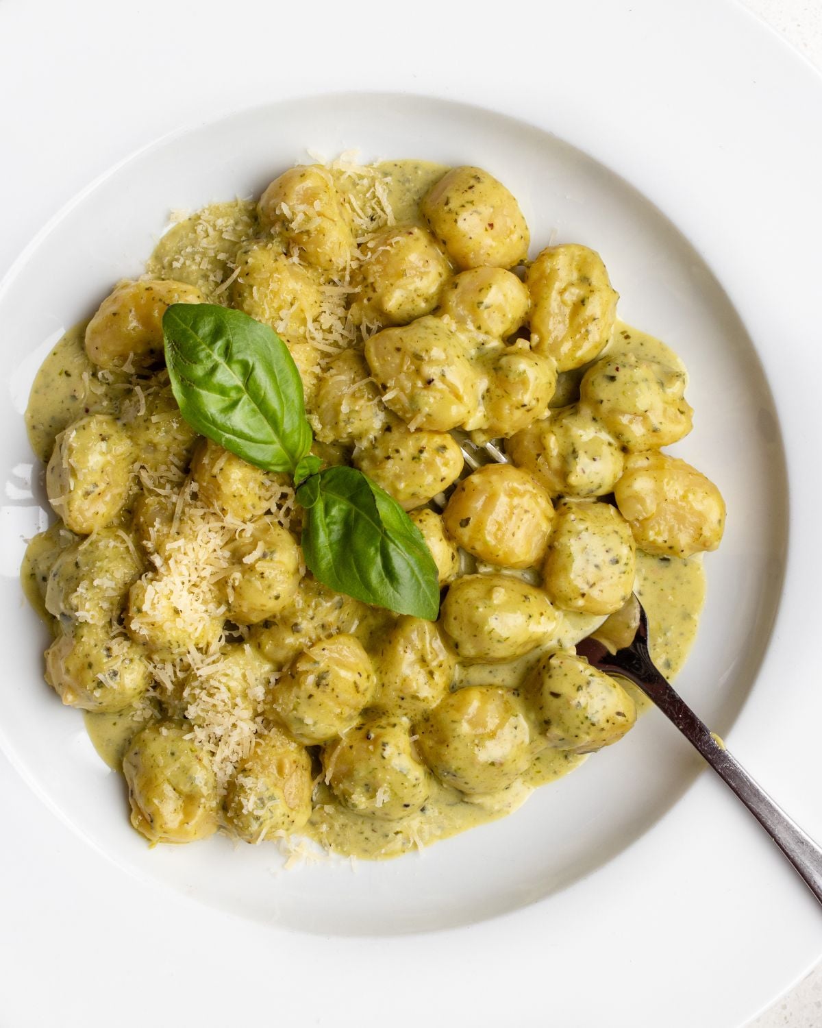 White plate with gnocchi in a creamy pesto sauce. Basil leaves on top and a fork resting in.