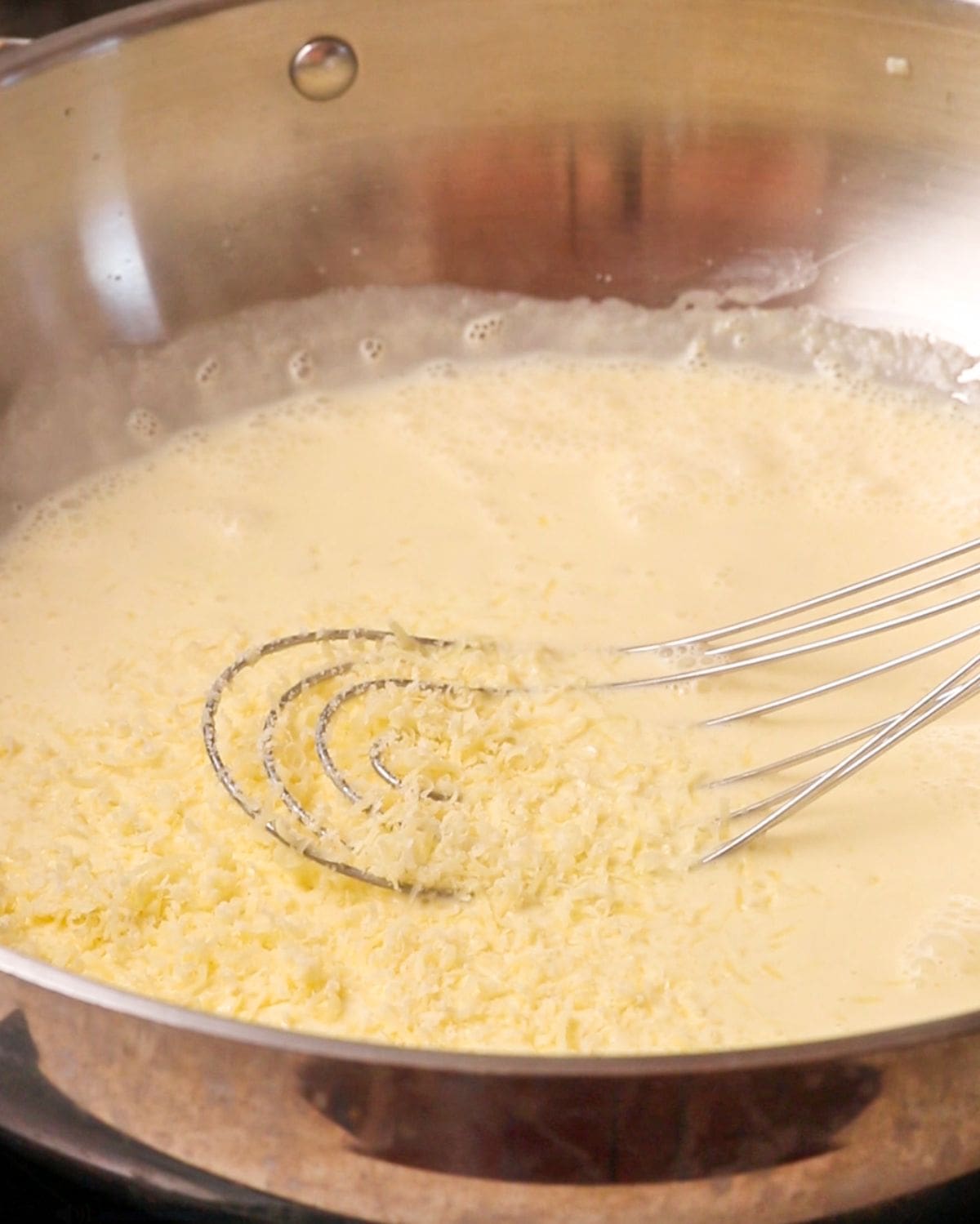 A whisk resting into cream in the pan with grated parmesan cheese sprinkled on top.