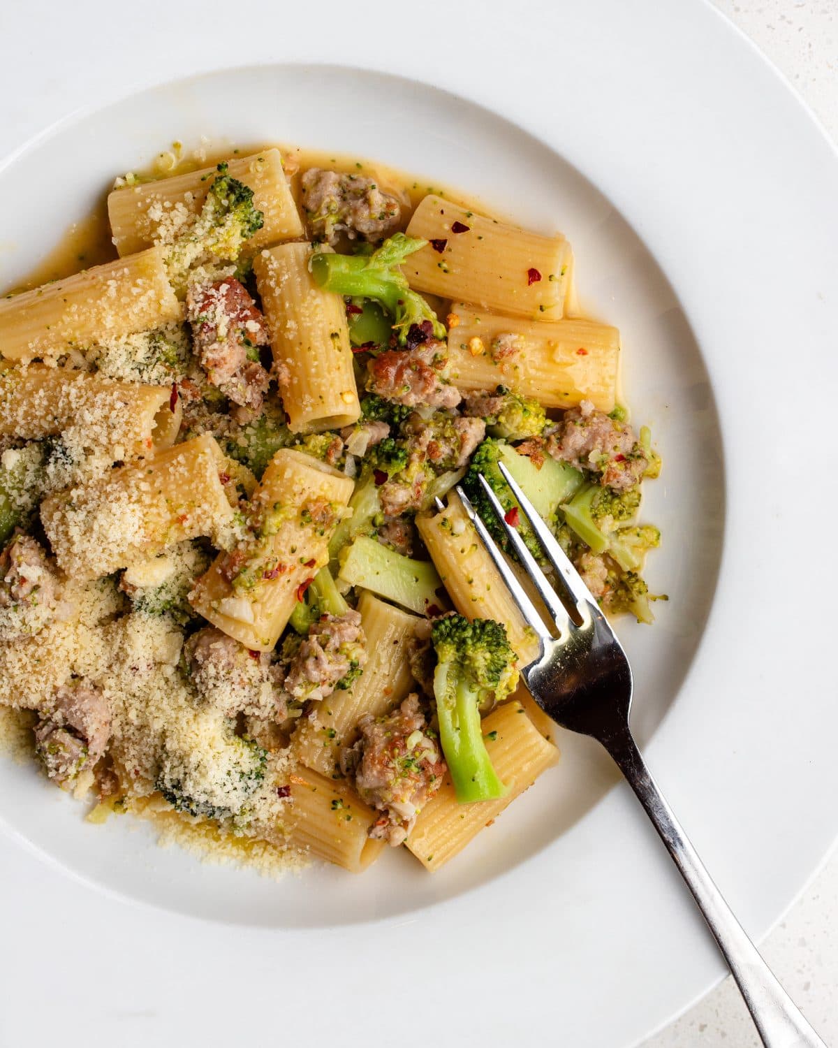 White plate with rigatoni pasta in a sausage and broccoli sauce. Parmesan sprinkled on top and a fork resting in.