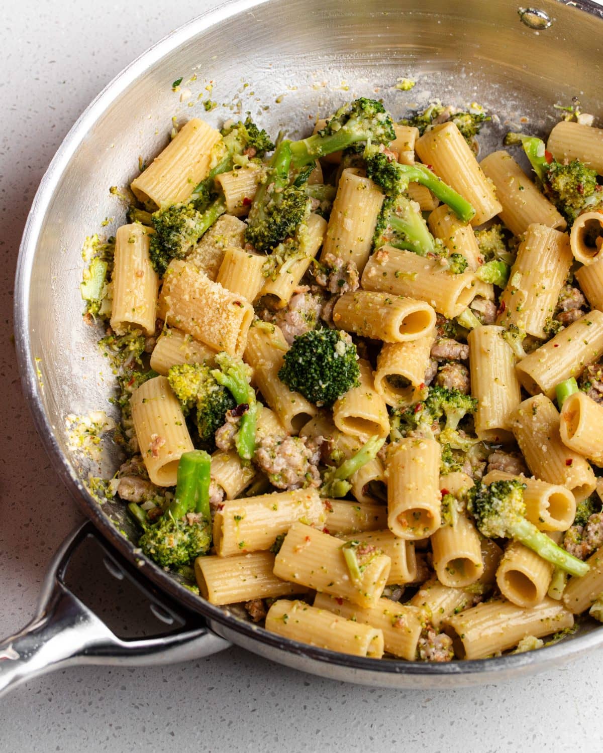 Stainless steel skillet on a bench top with rigatoni pasta in a broccoli and sausage sauce.