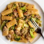 White plate with rigatoni pasta in a sausage and broccoli sauce. Fork resting in to pasta.