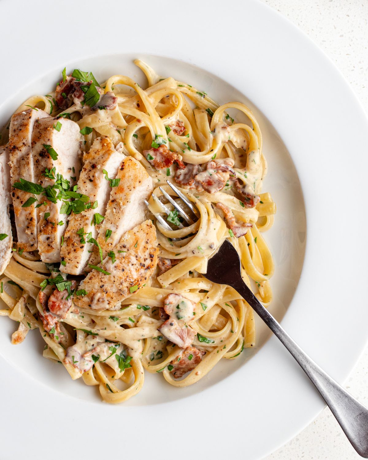 White plate with fettucine in a creamy sauce with bacon and chicken. Fork resting into pasta.