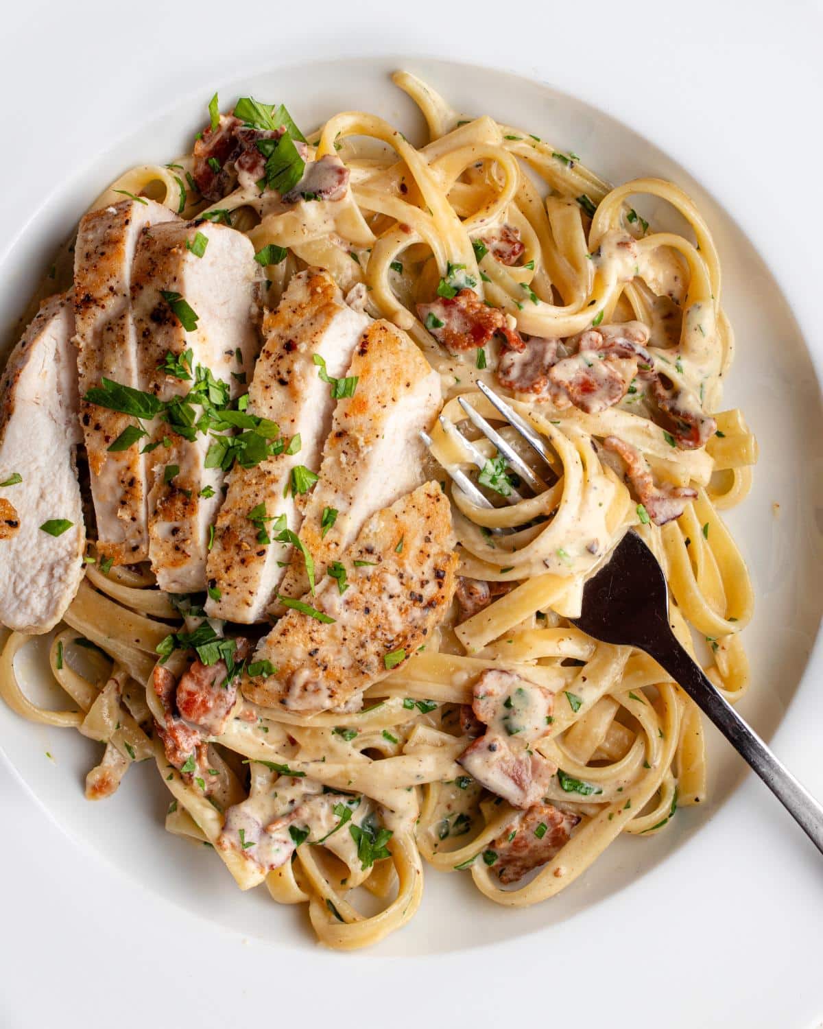 White plate with fettuccine pasta in a creamy bacon and chicken sauce. Fork resting in.