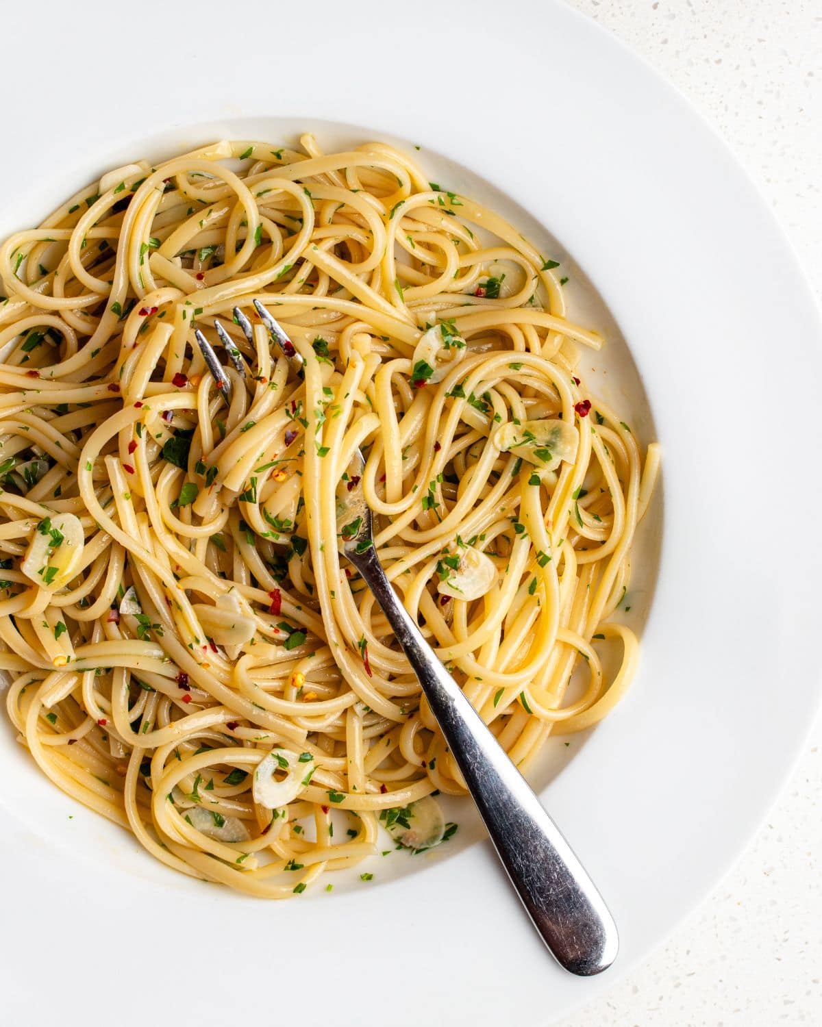 Spaghetti on a white plate in a aglio e olio sauce. Sliced garlic and parsley mixed through a a fork resting in.